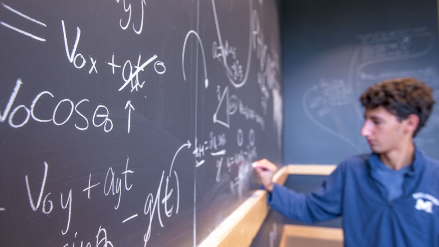 Student works on a math and science problem on a blackboard.